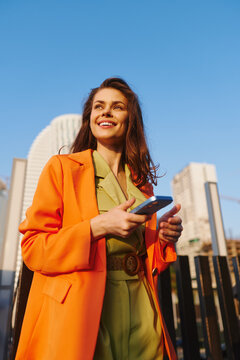 Smiling young woman in orange coat holds smartphone with both hands, texting outdoors against city skyline. Concept of messaging, digital connection and happy mobile communication.