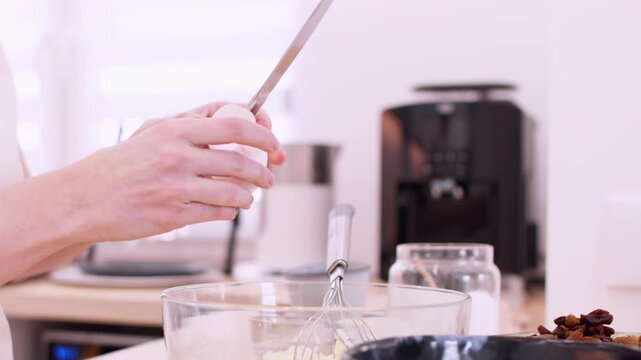 Homemade recipe. Preparing dough. Female chef hand cracking raw egg in glass bowl mixture with metal knife on modern kitchen table.
