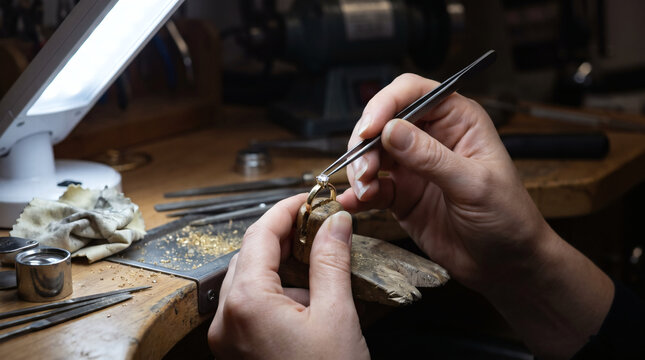 Professional jeweler hands using tweezers to set sparkling diamond onto gold ring at wooden workbench in jewelry making workshop