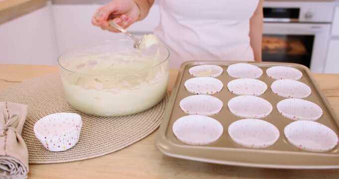 Preparing muffin. Treat recipe. Woman chef hand pouring batter from glass bowl spoon in paper liners in baking tin on home kitchen counter.