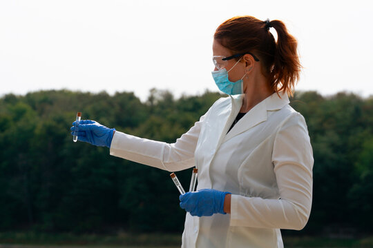 pneumonic plague outbreak scientist holding test tube sample, focused female epidemiologist in lab coat, face mask and blue gloves examining sample looking away from camera, forest and sky