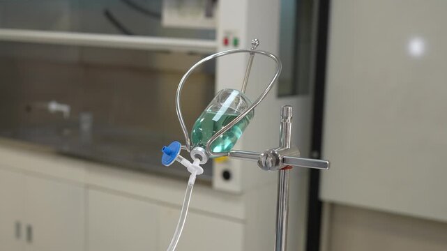 Gloved hand adjusting IV infusion set connected to inverted glass vial with blue solution on lab stand