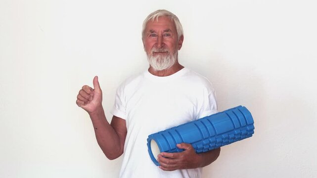 Senior man holding blue massage roller shows thumbs up on light background. Concept of approval and positive feedback for fitness gear, approval sign for sport store ads, social media and blog posts