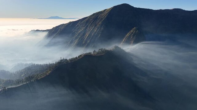 Mist curls around jagged mountain peaks. Blue sky frames the misty, silent summit near Bromo