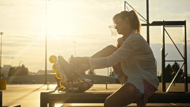 Young girl ties inline skates on bench at sunset. Blurry background shows active men playing basketball game on outdoor sports court during warm golden hour.