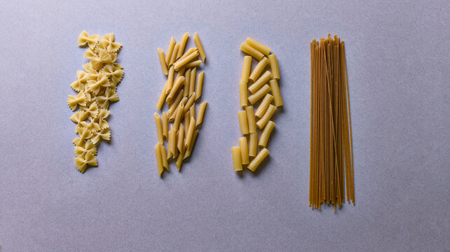 Dry pasta varieties sorted in vertical columns of farfalle penne rigatoni spaghetti on gray background.