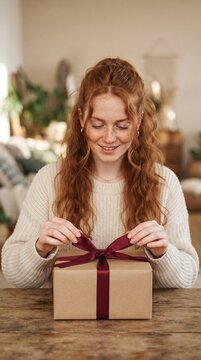 Young woman with red hair and freckles happily opening a gift box tied with a burgundy ribbon