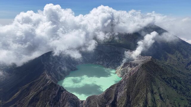 Vast clouds swirl above volcanic peak. Emerald lake rests in crater heart at Mount Ijen