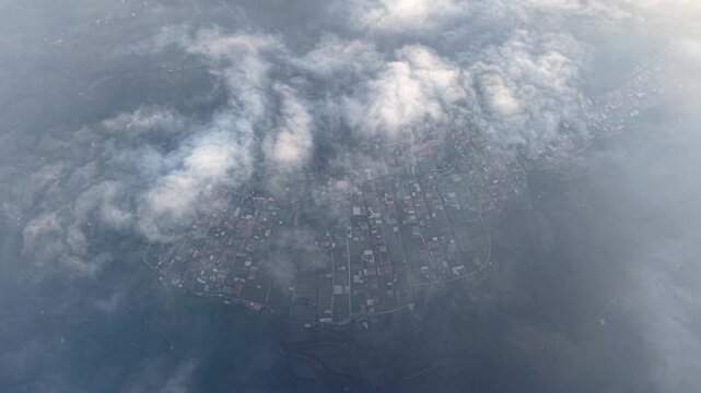 Clouds drift over a misty town below. Aerial view shows streets, buildings. The town appears quiet at Bromo