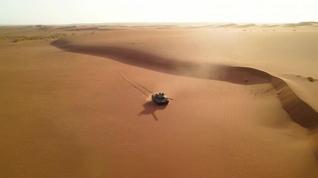 Tracked armored tank drives across broad orange sand dunes, leaving twin track marks and a light dust trail while warm sunlight casts a long shadow across open desert terrain