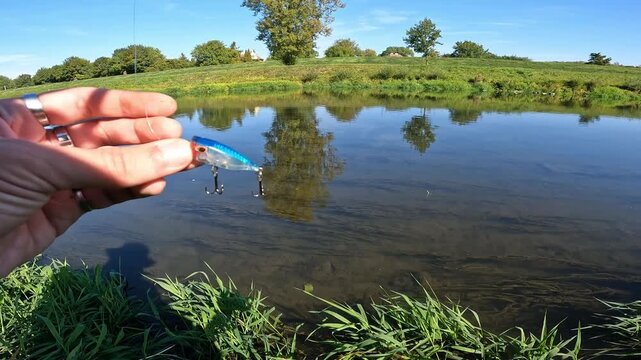 Angler holding blue and silver crankbait fishing lure with treble hooks before casting into river. Freshwater spinning tackle.