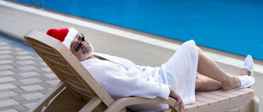 A happy adult man wearing a white robe and a Santa Claus hat, lounging by the pool. Relaxing hotel vacation during the New Year (or Christmas) holidays.