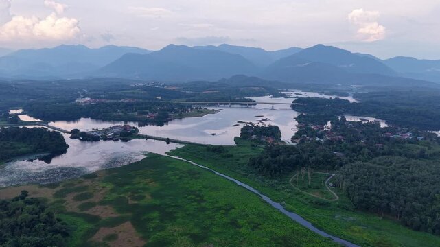 Aerial view showing winding river through green wetland with forest and mountain backdrop under a partly cloudy sky during daylight at Tasik Raban