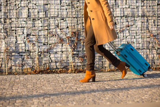 A woman in a long coat and brown ankle boots walks beside a stone wall, pulling a vibrant blue rolling suitcase under warm afternoon sunlight.