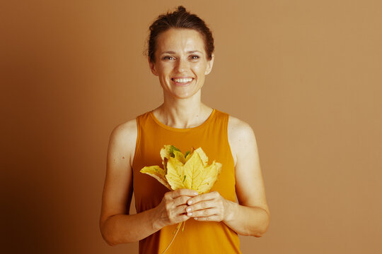 A cheerful woman with brown hair smiles brightly at the camera, holding a vibrant bouquet of yellow autumn leaves against a warm, plain brown background.