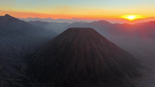 Aerial view of Gunung Batok rugged volcanic cone, layered mountains, clouds, and glowing sunset across Indonesia.