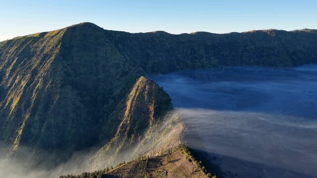 Misty dawn bathes volcanic crater lake in cool blue light. Rugged green slopes frame the still