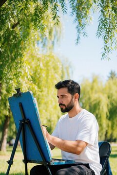 Young male artist creates caricature drawing on blue easel in sunny park. Creative man sits outdoors sketching portrait on paper. Talented person practices art hobby during summer leisure time under