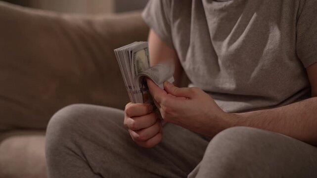 Man counting, flipping through cash while sitting on a sofa at home. Hands hold a thick stack of US dollars banknotes. Money, income, savings, salary and personal finance concept. Authentic lifestyle