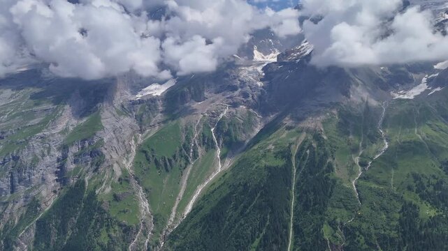 Drone perspective of dramatic green alpine valleys, rocky cliffs, snow patches and low clouds in the Swiss mountains during summer.