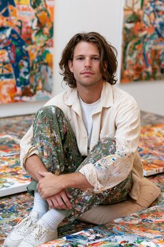 Young male artist sitting among colorful abstract paintings in a creative studio, wearing paint-splattered clothes and looking at the camera, vertical lifestyle portrait