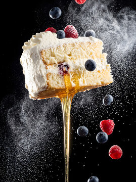 Slice of vanilla berry sponge cake with dripping honey and falling fresh blueberries and raspberries on a dark black background