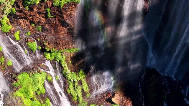 Mist rises from a powerful waterfall. Green moss clings to dark, wet rock faces at Tumpak Sewu