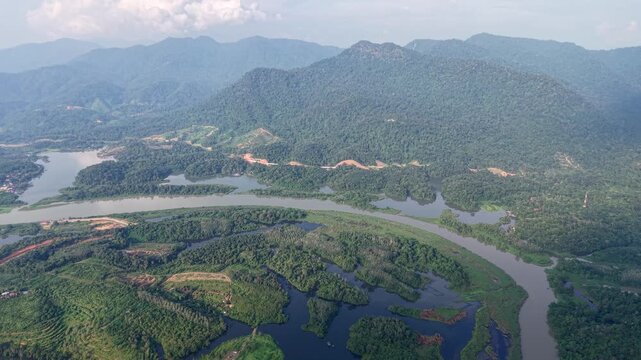Aerial shot of a wide river winding through dense green tropical forest and mountain landscape under a partly cloudy sky during daylight at Tasik Raban