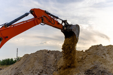An orange hydraulic excavator bucket releases soil onto a large earth pile at a construction site, with the boom and hydraulic cylinders visible beneath a cloudy sky. © Marina