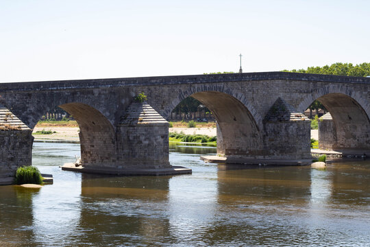 Stone arch bridge spanning a river with reflections visible in the water, surrounded by greenery and clear blue sky on a sunny day