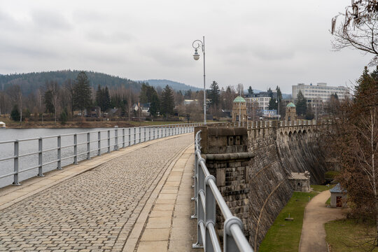 Mseno Reservoir dam walkway in Jablonec nad Nisou, Liberec Region, Czech Republic, with stone paving, railings, water and wooded hills beyond.