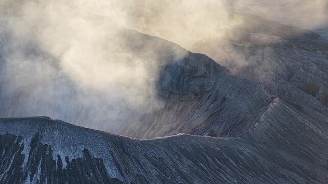 Aerial sunset over Mount Bromo crater, mist drifting across serrated ridges with tiny hikers