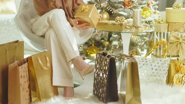 A woman in an elegant armchair carefully examines a gold gift box, surrounded by a pile of luxurious presents and a festive Christmas tree, creating a lavish holiday scene.