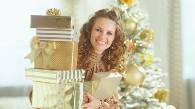 A woman with curly brown hair smiles joyfully at the camera while holding a large stack of gold and white Christmas presents. A blurred, decorated Christmas tree creates a bright, festive background.