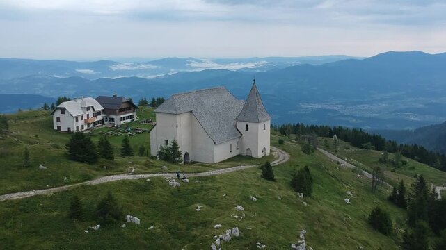 Scenic drone shot of Saint Ursula Church and mountain hut atop Urslja gora in Slovenia, surrounded by grassy alpine ridges with expansive views of valley mist and distant mountains.
