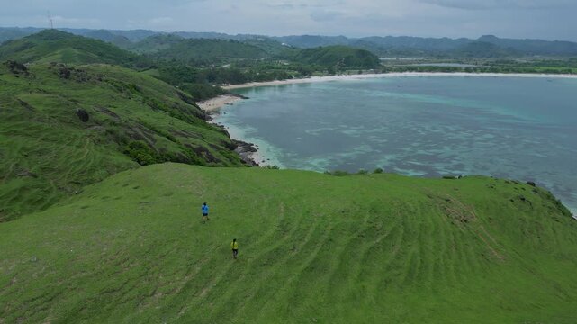 Explorers Standing At Cliff Edge Overlooking Turquoise Bay And Curved Beach, Aerial View Shows Reef Patterns In Shallow Water, Green Hillside Foreground, Contemplative Lookout