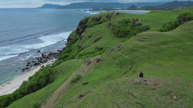 Woman Walking Along Coastal Hilltop Path, Sweeping Bay And Turquoise Water In Background, Panoramic Aerial View Of Rounded Green Knoll, Relaxed Pace, Leisure Travel And Nature, Lombok island,Indonesia