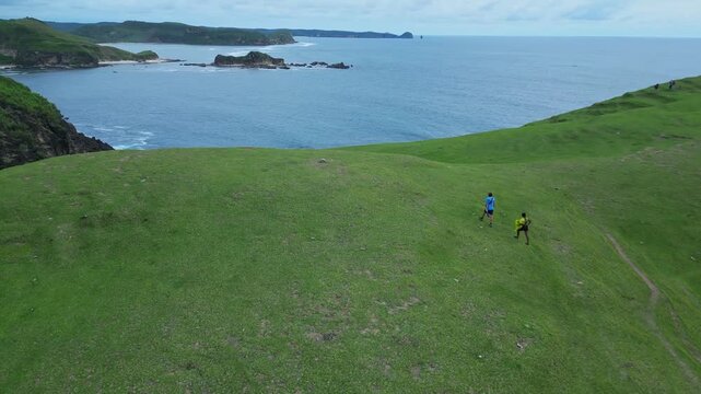 Couple Hiking Along Coastal Ridge Aerial Drone View Showing Two Hikers In Blue And Yellow Jackets Crossing Green Grassy Hill Above Ocean And Rocky Islets, Overcast Sky, Peaceful Travel Mood,Lombok