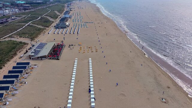 High-angle aerial drone clip of broad sandy beach, North Sea shore, beach cabins, resort pavilions, and coastal dunes in Katwijk aan Zee, South Holland, Netherlands on bright summer day