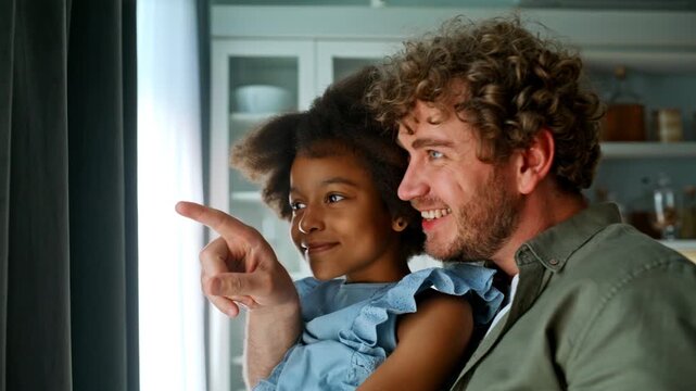 Multiracial dad holding daughter pointing window. Closeup smiling girl home.