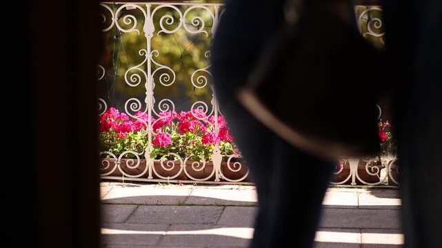 View through a doorway of an ornate white balcony railing with vivid pink geraniums, partially obscured by two silhouetted figures walking past in the foreground.