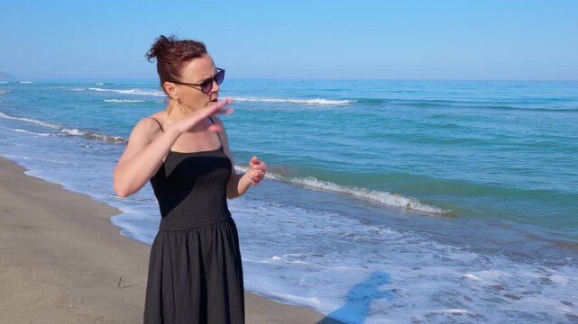 Woman in a black dress feeling unwell on a hot beach. Overheated caucasian woman in a black dress standing on a sunny beach and feeling dizzy due to the high temperature, she is fanning herself