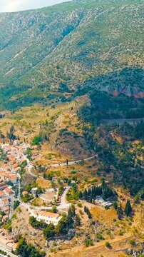 Vertical video. Delphi, Greece. Drone footage of ancient theater and Temple of Apollo ruins on Mount Parnassus slopes, modern town with terracotta rooftops in the valley below, bright summer sun. Dro