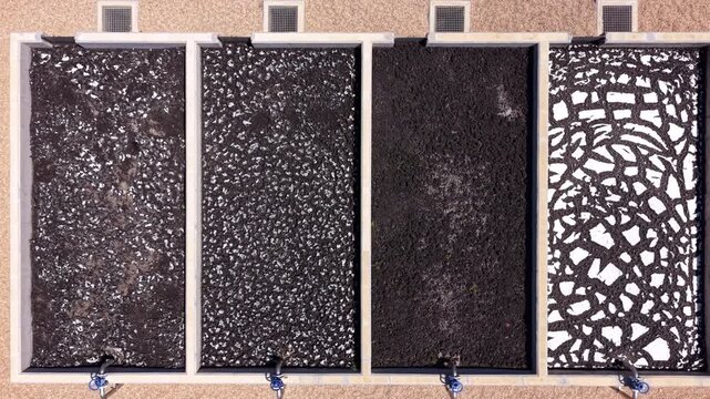 Overhead view of four sludge drying beds displaying sequential drying stages from dark wet sludge to fully dried cracked biosolids with white cracked mosaic pattern at wastewater treatment facility