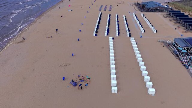 High-angle aerial drone clip of organized beach cabins, North Sea shoreline waves, resort pavilion, and sandy coastline in Katwijk aan Zee, South Holland, Netherlands on bright summer day