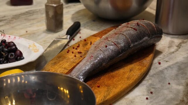 Close up view of chef seasoning raw whole fish on wooden cutting board with pink peppercorns in kitchen setting with bowl of cherries and sharp knife nearby on marble countertop.