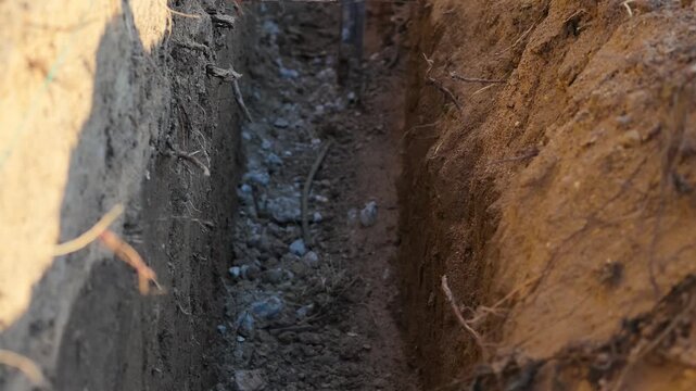 Close-up view of narrow, freshly dug trench in earth, revealing soil, rocks, and plant roots along sides.