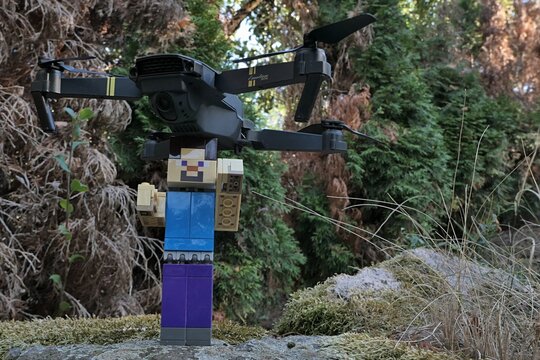 Minecraft action figure of smiling main characters Steve standing on moss covered garden stone slab with budget drone quadcopter Eachine E58 on his head. Thuja trees in background. 