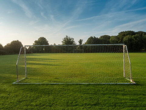 White soccer goal stands alone on green grass. Trees line the background under blue sky with wispy clouds. Scene peaceful and quiet, no people present. Ideal for sports, recreation