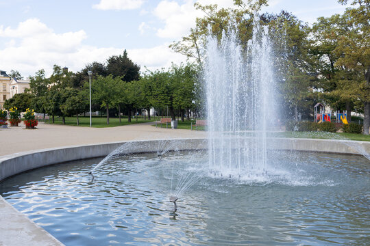 Urban park fountain with tall water jets, circular basin, trees, pathways, historic buildings, and playground in a landscaped public garden